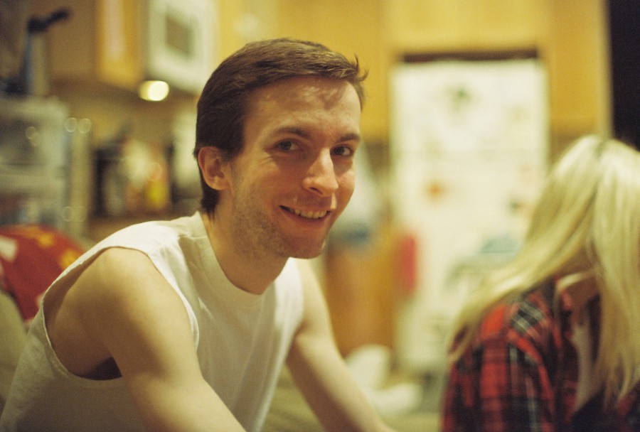 A warm, candid indoor portrait shot on 35mm film with a vintage Petri SLR, showing Ethan (me!) sitting on a couch inside a West Town apartment on the Fourth of July, 2013. I'm wearing a sleeveless white tank top and smiling directly at the camera. My short brown hair is gently tousled, and the golden film lighting softens my features. In the background, slightly out of focus, Lauren sits beside me, turned away in conversation with someone off-frame. Her long blonde hair (the blonde side of her split-dye) is visible, and she wears a red plaid shirt. Behind us, a refrigerator with magnets and a wood-paneled kitchen add cozy context to the holiday hangout. Caitlin took this photo.