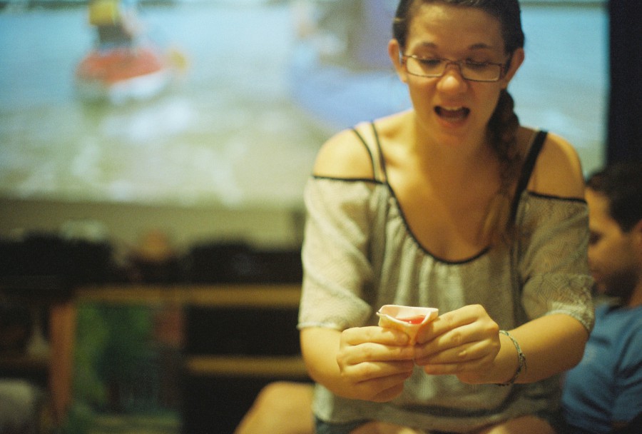 A candid and energetic photo taken indoors on 35mm film, showing Amanda at the center of the frame, holding a Jello shot in both hands and reacting dramatically. She wears a loose, semi-sheer black-and-white off-the-shoulder top, glasses, and has her hair pulled into a braid over one shoulder. Behind her, Justyn lounges casually in a blue shirt, partially visible. The far wall is covered by a large projected image—from a home projector—showing a boating or water scene, adding a fun backdrop to the party moment. The film grain adds a charming softness to the motion and expression.