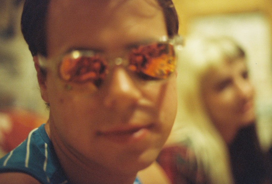 A tight 35mm film close-up of Elliott indoors, wearing reflective, mirrored sunglasses with orange-red tints that dominate the photo. The reflections are chaotic and vivid, distorting the room and making his gaze mysterious. Elliott is slightly out of focus, adding a dreamy or dazed quality, and wears a blue tank top with white stripes. In the background, Lauren is blurred but visible, her blonde hair catching the warm light. The composition is tight and slightly chaotic, making it feel like a spontaneous flash of memory during a rowdy holiday evening.