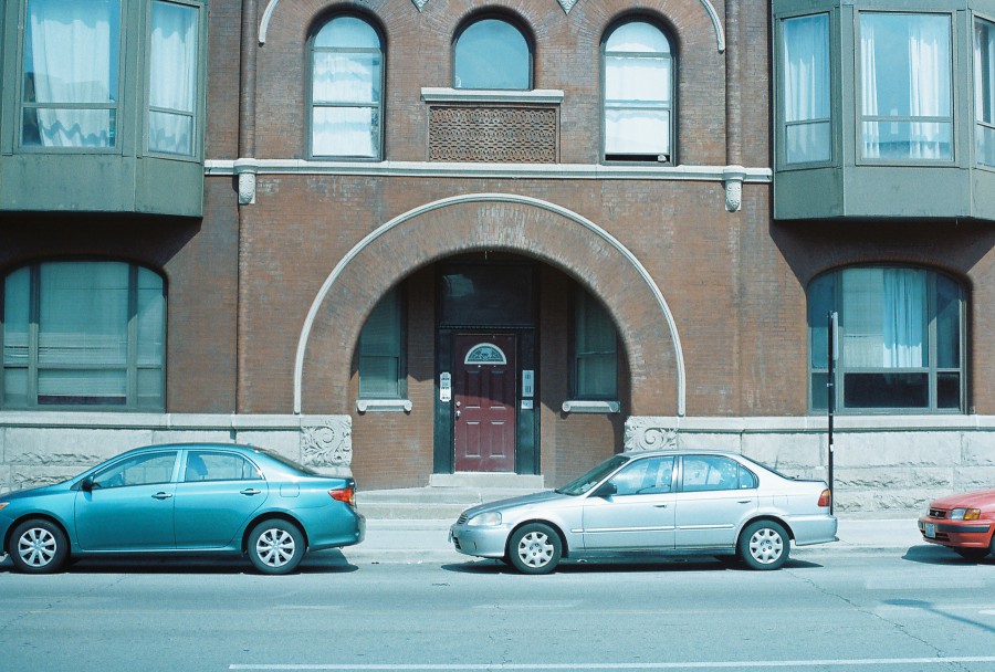 A daytime 35mm film photo of the front façade of 917 S Racine Avenue in the Little Italy neighborhood of Chicago. The building is a classic Chicago brownstone-style apartment with a large central arched entranceway framed by light stonework. The door is deep red with a small semicircular window at the top, flanked by intercoms. Above the arch are three tall windows, and additional bay windows on both sides jut outward with large panes and curtains visible inside. Parked in front are three cars—a teal Toyota Corolla, a silver Honda Civic, and a red sedan—lined up on the gray asphalt of the street. The sunlight is harsh and direct, creating strong shadows and giving the image a punchy blue cast from the film stock.