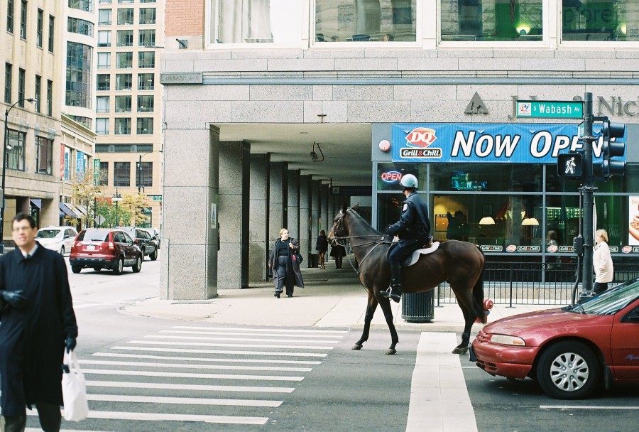 A busy city intersection at Wabash Avenue and Congress Parkway (now Ida B. Wells Drive), featuring a police officer on horseback in the crosswalk. The "DQ" Dairy Queen Grill & Chill storefront in the background prominently displays a "NOW OPEN" sign. Pedestrians move along the sidewalks, and a mix of cars, including a red sedan and a SUV, pass by. The modern urban architecture blends with classic Chicago charm, with clean, crisp lines in the shot captured on 35mm film, giving it a warm and slightly vintage aesthetic.