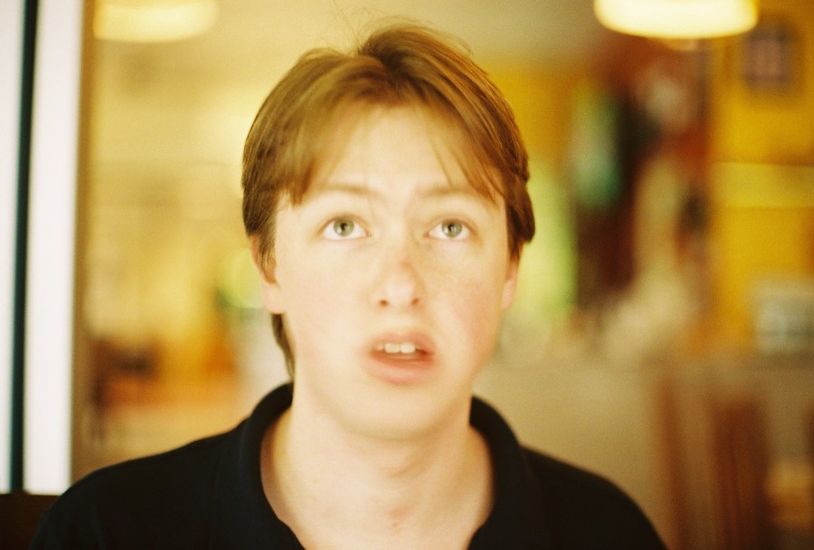 Another candid shot inside Thai Spoon restaurant on Wabash and Harrison, featuring Andrew again. He has a vacant, confused expression, his mouth slightly open as if mid-sentence or lost in thought. The soft focus creates a dreamy, almost surreal effect, with the restaurant's warm, golden lighting gently illuminating his face. The blurry background adds to the sense of relaxed ambiance, typical of a laid-back meal with friends. Taken on 35mm film with a Petri camera, emphasizing the candid, unguarded moment.