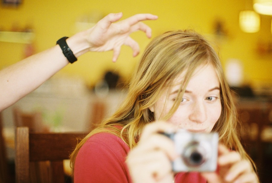 A candid, close-up shot of Andria sitting in Thai Spoon, a restaurant with a warm, yellow interior. She has long, flowing blonde hair that partially obscures her face, wearing a bright pink sweatshirt. She is holding a silver Canon Powershot camera up to her face, as if about to take a photo, and looking playfully to the side with a slight smirk. Behind her, a hand with a black wristwatch reaches into the frame from the left, making a claw-like gesture toward her head, adding a playful and spontaneous feel to the shot. (It's Andrew.) The background is softly blurred with yellow walls, wooden chairs, and warm ambient lighting, creating a cozy and casual atmosphere. Captured on a Petri 35mm film camera, the image has a soft, almost dreamy quality, typical of film photography.
