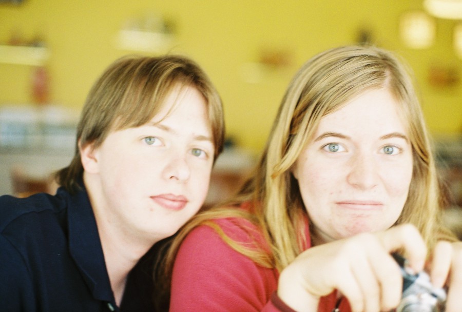 A casual portrait of Andria and Andrew sitting together in Thai Spoon, a small restaurant with a warm yellow interior. Andria, on the right, has long, wavy blonde hair and is wearing a bright pink sweatshirt, holding a small silver Canon Powershot camera. Her expression is a mixture of surprise and amusement, eyes wide and eyebrows raised. Andrew, on the left, has light brown hair and is wearing a dark navy blue polo shirt. He is leaning slightly into the frame, looking calm and neutral with a subtle, relaxed smile. The shallow depth of field gives the image a dreamy, vintage feel, with the background of wooden chairs and soft, diffused light appearing slightly blurred. The atmosphere feels friendly and lighthearted, with the image capturing a spontaneous, candid moment between friends.