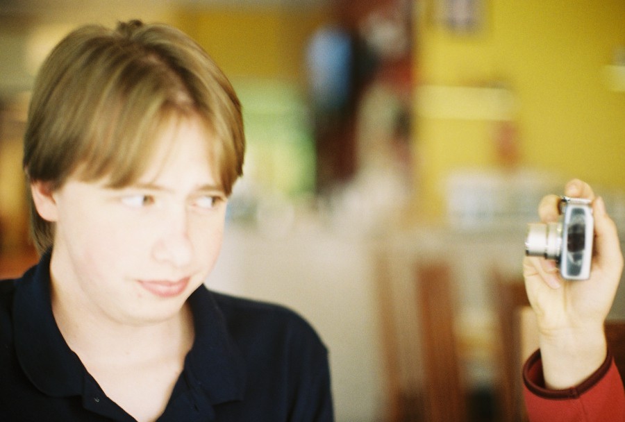 A close-up shot of Andrew sitting at a table inside Thai Spoon, looking off to the side with a skeptical and slightly annoyed expression. He has light brown hair parted down the middle and is wearing a dark navy blue polo shirt. His face appears softly illuminated by the warm ambient light of the restaurant, which features yellow walls and a cozy, slightly cluttered interior. In the foreground, Andria’s hand holding a silver Canon Powershot camera enters the frame from the right, pointing it toward Andrew, adding a sense of playful interaction. The focus is slightly soft, typical of film photography on a Petri 35mm camera, creating a gentle and nostalgic aesthetic.