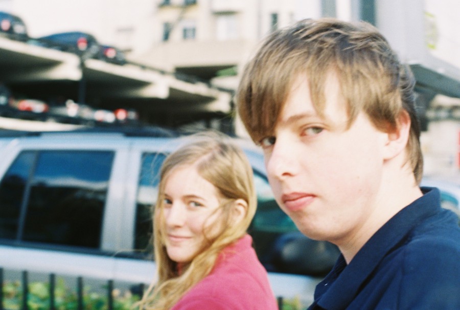 An outdoor shot of Andrew and Andria walking along a city sidewalk. Andrew is in the foreground, wearing a dark navy blue polo shirt, with his head turned slightly to the side, looking back at the camera with a neutral, almost deadpan expression. Andria walks a little further back, smiling warmly with long, wavy blonde hair, wearing a bright pink sweatshirt. The background is bustling with urban details, including parked cars and a multi-level parking garage. The lighting is natural and bright, giving the image a candid, spontaneous feel, typical of a day out in Chicago’s South Loop area. The colors are vibrant and slightly washed out, capturing a casual city moment.