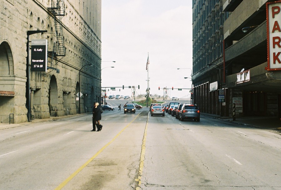 A photo looking east down Congress Parkway (now Ida B. Wells Drive) in Chicago, showing a divided roadway with faded yellow lane markings and light traffic moving toward the horizon. On the left, the imposing stone architecture of Roosevelt University’s Auditorium Building looms, with a prominent fire escape visible. A large sign advertising an R. Kelly performance on November 17 and 18 hangs from the building (a detail now recognized with discomfort). On the right, a multi-level parking garage displays a large red "Park" sign. A pedestrian dressed in dark clothing crosses the road in the mid-ground, and the sky appears partly cloudy with diffused sunlight. The perspective emphasizes the vastness of the street and the contrast between old stone architecture and modern infrastructure.
