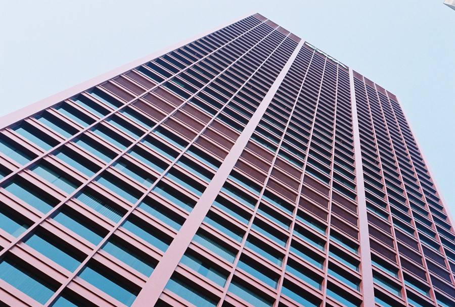 A dramatic upward perspective shot of the CNA Center, a bright red high-rise building in Chicago’s South Loop. The building’s modern grid-like facade of windows and red-painted concrete stretches vertically into the sky, giving a towering, monolithic impression. The sky above is pale and cloudless, creating a stark contrast with the deep, bold red of the building. The shot emphasizes symmetry and repetition in the architecture, capturing the structure’s immense scale and geometric design. The photo’s sharp, clean lines and vivid colors create a bold, almost abstract aesthetic, highlighting the building’s iconic presence in the Chicago skyline. The image has a crisp, clear quality, typical of Petri 35mm film photography, showcasing the architectural details with striking precision.