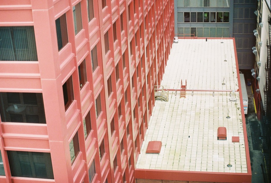 The image shows a downward perspective of the CNA Building in Chicago, taken from a higher vantage point, a dorm room at Columbia College Chicago. The iconic red exterior of the CNA Building is prominent, with rows of evenly spaced windows. The adjacent rooftop of a shorter section of the building is visible in the foreground, covered with a flat, light-colored surface and featuring red vent structures. The urban canyon effect is created by tall buildings on either side, and the light, overcast sky adds a subtle blue hue to the ambient lighting. Captured with a Petri 35mm film camera on May 1, 2010.