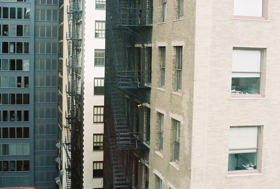 An urban view from a dorm building in the South Loop of Chicago, showing a complex arrangement of fire escapes and narrow alleyways between tall structures. The central building features classic brickwork with old-fashioned fire escapes stretching vertically along the façade, while neighboring modern buildings showcase sleek, dark metal and glass exteriors. The shot captures the mix of historic and modern architecture typical of the area. Subtle reflections can be seen in the windows, hinting at the urban density. Taken with a Petri 35mm film camera, the image has a slightly muted, nostalgic tone.