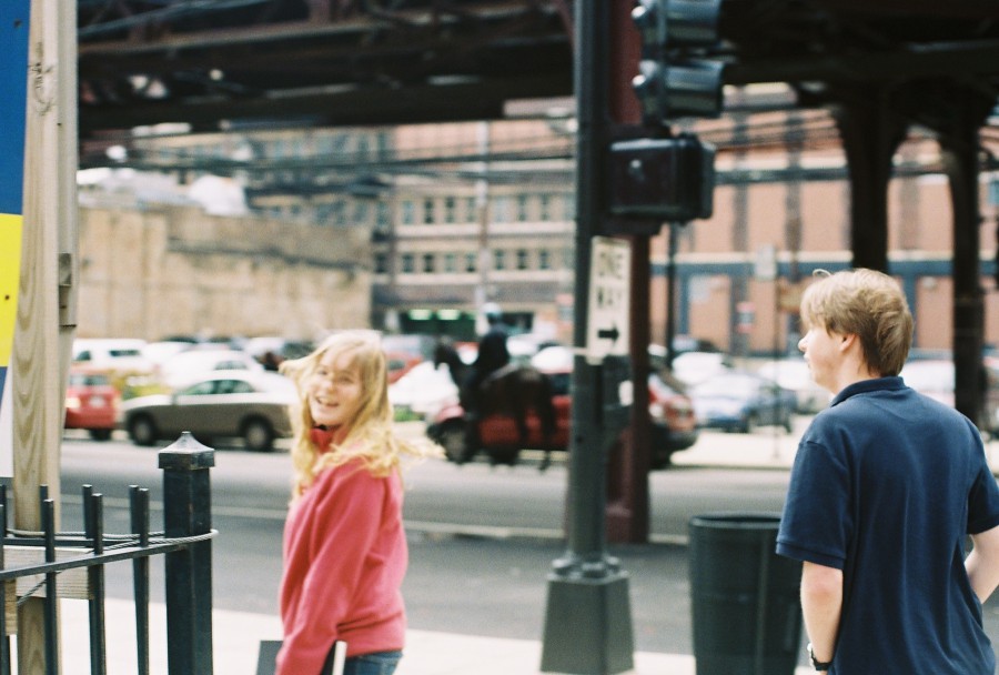 A dynamic street scene at the intersection of Wabash and Van Buren in Chicago. Andria, wearing a vibrant pink jacket, is walking on the left, glancing back with a cheerful expression, her long blonde hair catching the light. Andrew, in a dark navy polo shirt, walks to the right. A mounted police officer on a horse is seen in the background, partially obscured and blurred, while the iconic elevated "L" train tracks loom above. Cars fill the parking lot behind the group, and various urban details, like street signs and fences, add depth to the scene. Shot on a Petri 35mm film camera, capturing a sense of movement and spontaneity.