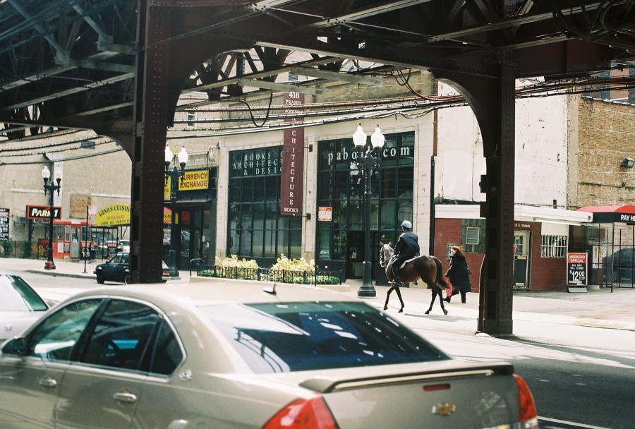 The photograph shows an elevated train track ("L" tracks) running along Wabash Avenue in downtown Chicago. Under the tracks, a mounted police officer on horseback patrols the street, giving a unique urban-cowboy juxtaposition. On the right, the storefront of "Prairie Avenue Bookshop" with a sign advertising "Books on Architecture & Design" can be seen, along with a Currency Exchange shop nearby. A mix of parked and moving cars adds to the bustling atmosphere. The photo, taken with a Petri 35mm film camera, features soft, natural lighting.