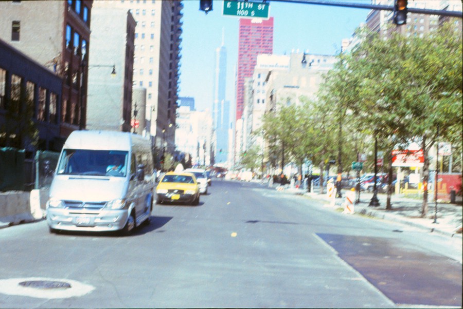 A slightly blurred photograph of the intersection at 11th Street in Chicago, taken in 2009 on 35mm slide film. The red CNA building stands prominently in the distance, surrounded by a cluster of other downtown structures. Trees line the right side of the street, while a silver van and a yellow taxi are visible driving along the road. Orange and white construction barriers mark the edges of the sidewalk. The motion blur adds a dynamic yet hazy quality to the image, typical of film photography.