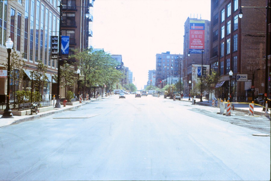 A photograph looking down a street in the opposite direction, showing symmetrical buildings on either side with large glass windows. The left side of the frame features trees and neatly landscaped areas, with parked cars and a lamppost visible. On the right, tall brick and glass buildings rise into the sky, with banners promoting a "landmark on the rise" development. The wide, open street includes minimal traffic, with light construction cones lining the sidewalks.