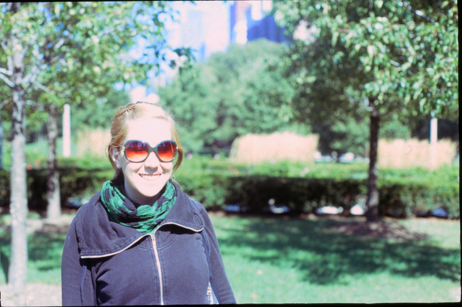 A portrait of Chelsea standing in a sunlit green park. She wears a zipped-up black hoodie with a dark green scarf wrapped loosely around her neck. Oversized, round sunglasses reflect the bright sunlight, adding a vibrant touch to her casual look. The park is dotted with small trees and neatly trimmed grass, while blurred foliage and tall buildings in the distance provide a layered backdrop. The image is slightly overexposed, giving it a soft, airy feel.