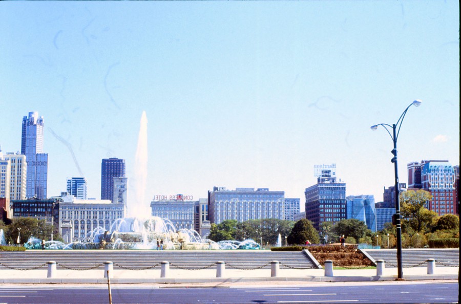 A stunning wide shot of Buckingham Fountain in Chicago, with its cascading white water reaching into the air against a backdrop of iconic city buildings. The Congress Hotel and other historic structures can be seen in the distance, framed by a clear blue sky. The foreground includes a chain barricade and stone steps leading up to the fountain. The image captures both the grandeur of the fountain and the architectural beauty of the surrounding area, exemplifying the charm of downtown Chicago.