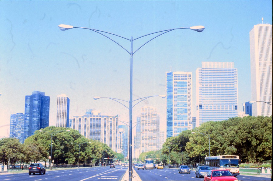 A symmetrical street-level view down the center of a divided highway in Chicago. Tall streetlights on either side frame the image, converging toward a vanishing point in the distance. Cars, a bus, and taxis populate the road, while dense greenery lines the sidewalks on both sides. Skyscrapers of varying designs rise in the background, creating a striking cityscape. The film's fuzz and minor imperfections lend a nostalgic texture to the shot.