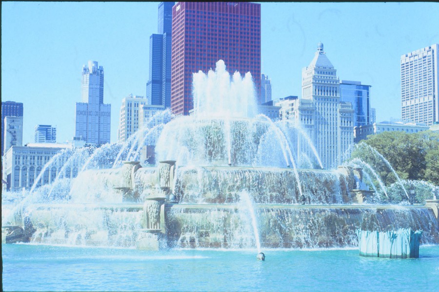 A close-up professional 35mm film highly-detailed shot of Buckingham Fountain, highlighting its intricate stone carvings and powerful streams of water. The red CNA building looms directly behind the fountain, alongside other skyscrapers that create a dramatic backdrop. The bright blue sky and reflections in the water enhance the photo's clarity, making it a perfect representation of Chicago's architectural beauty and vibrant atmosphere.