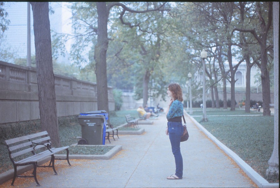 A candid portrait of Amanda standing in Grant Park, framed perfectly by the vanishing point. She faces sideways, wearing fitted jeans, a patterned blue top, and a brown shoulder bag. The park features long benches, green trash cans, and a paved path lined with trees. In the distance, a man sits on a bench, and the arched windows of the Art Institute of Chicago peek out from the background on the right. The soft, overcast lighting creates a serene, balanced composition.