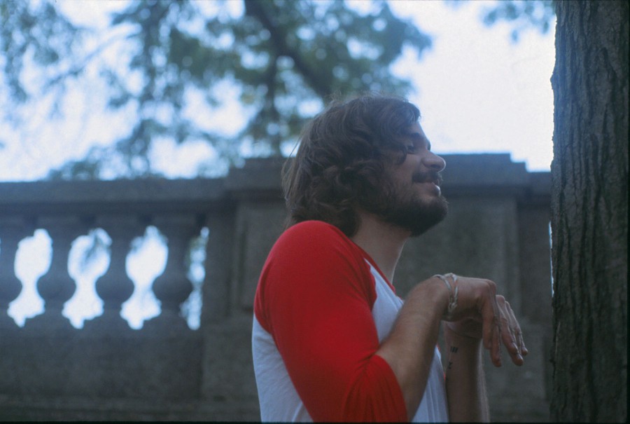 A portrait of Eric in Grant Park shows him with a playful expression, making a funny gesture with his hands as he stands next to a large tree. His red and white baseball tee contrasts with the soft green leaves in the background. Behind him, a stone balustrade hints at the park's classical design. The image captures a candid moment, with natural light illuminating his hair and beard.