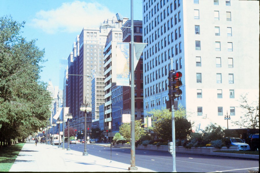 A cityscape from the South Loop area of Chicago features a bustling sidewalk along Grant Park, flanked by tall, iconic Chicago buildings. The scene is alive with details, from banners hanging on streetlights to the greenery of trees lining the street. The clean, bright sky adds contrast to the sharp architectural lines captured with the film’s characteristic vibrancy.