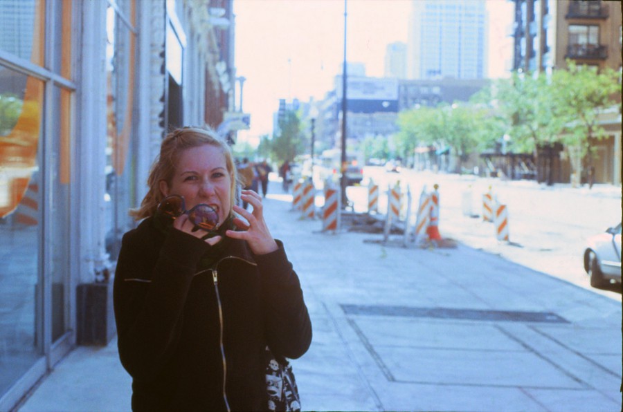 Chelsea poses humorously on a sunlit sidewalk, holding sunglasses and making a goofy face with exaggerated hand gestures. She wears a black jacket, and her hair is pulled back. To her left, the Columbia College building reflects the sunlight, while a series of orange and white construction cones lines the sidewalk. The background features distant trees and light urban traffic.