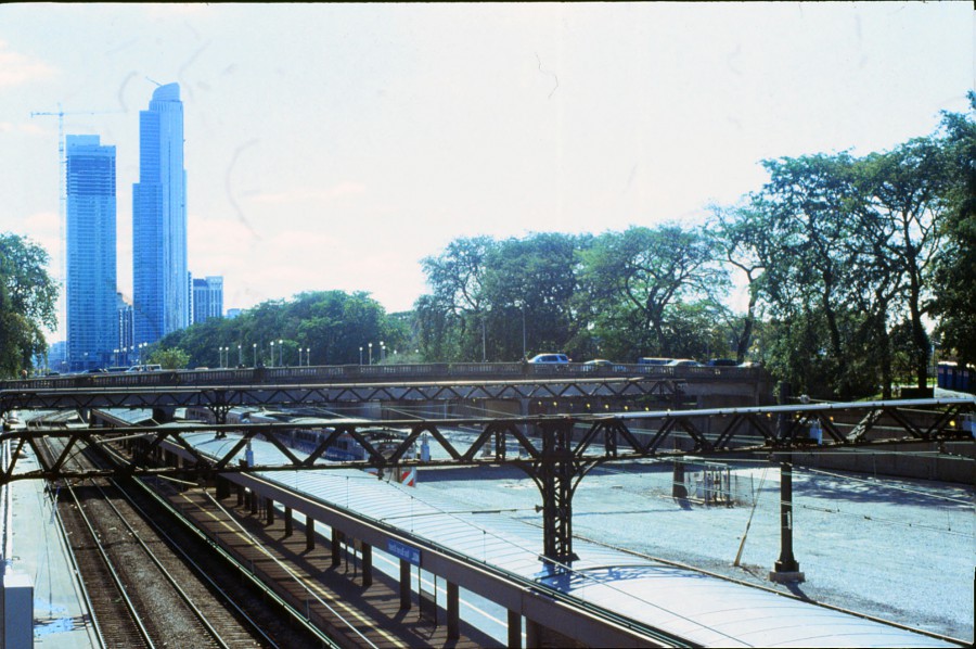 A train station shows the tracks disappearing into the distance, with tall skyscrapers rising behind a canopy of trees. A pedestrian bridge stretches over the tracks, its metallic structure casting shadows below. The slide film captures the urban energy and sharp details of the Chicago skyline, with a construction crane visible among the buildings.