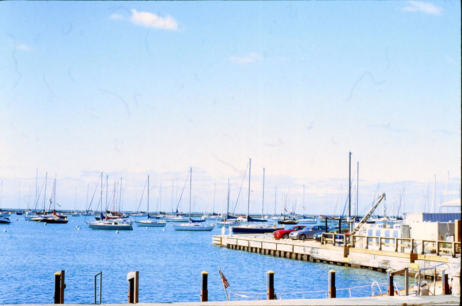 A harbor scene focuses on a wooden dock and a few parked cars, with rows of sailboats floating on the calm blue waters of Lake Michigan in the background. The dock's weathered planks and mooring posts frame the image, while the clear sky provides a serene contrast to the bustling activity of the harbor.