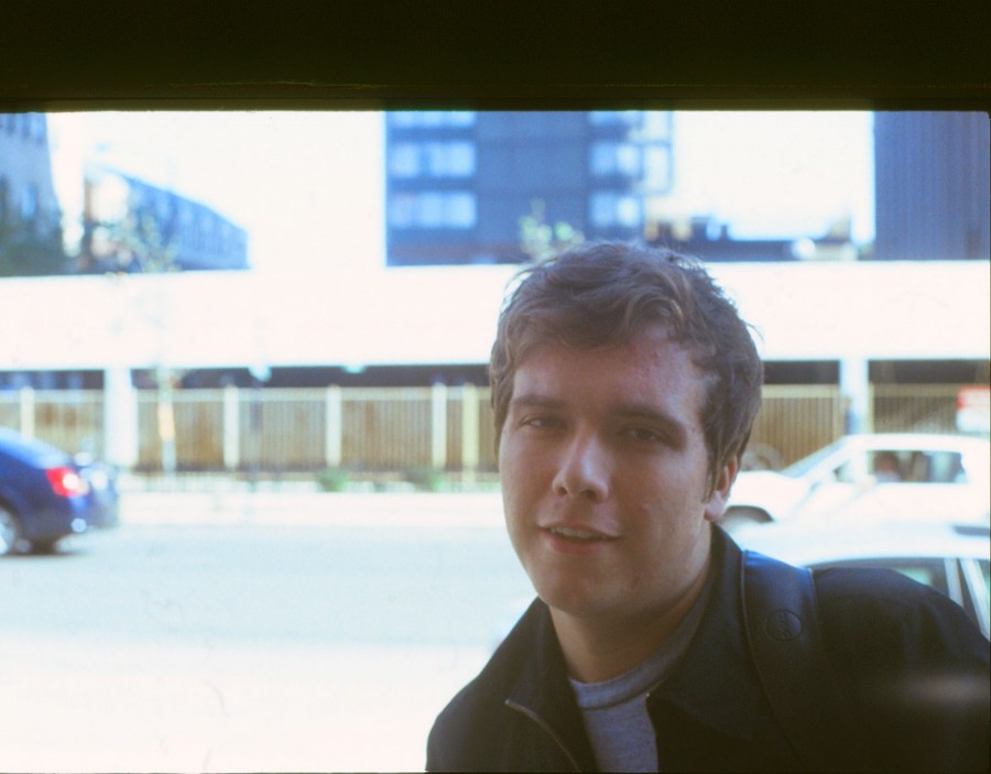 A close-up portrait of Tony captures him wearing a jacket and the strap of a backpack over his shoulder. He gazes directly into the camera with a casual expression, framed by blurred urban elements in the background. The bright light and soft focus bring a nostalgic quality to this moment, typical of 35mm slide film photography.
