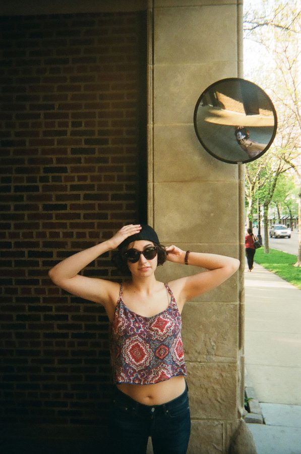 Katie adjusts her black cap with both hands while wearing dark sunglasses. She stands near a stone column with a circular traffic fisheye mirror mounted above, reflecting her slightly distorted image along with some greenery and the sidewalk in the background. She wears the same red and blue patterned crop top and dark jeans. The mirrored reflection adds a quirky touch to the film photograph.