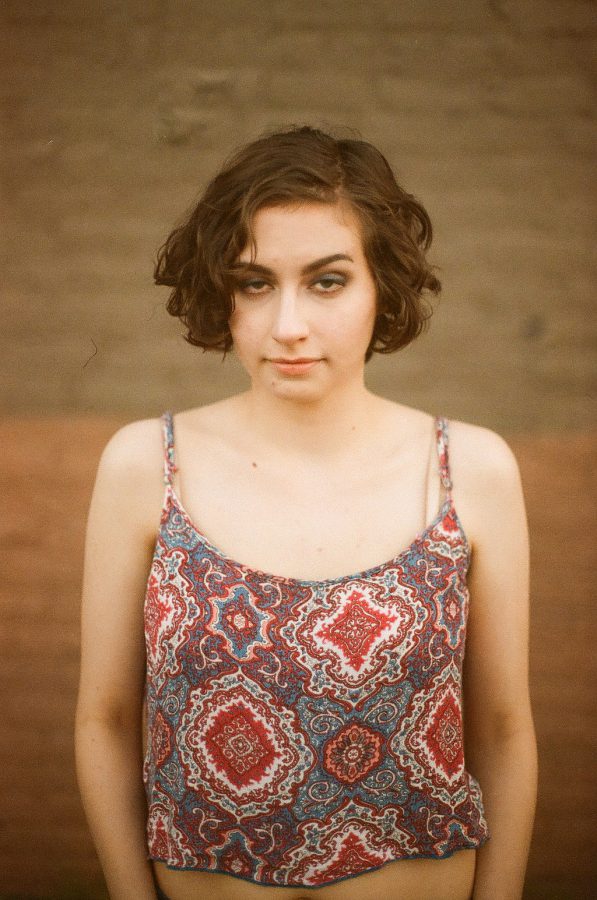 Katie stands against a blurred brick wall, her short curly brown hair framing her face. She wears a red and blue patterned sleeveless crop top with intricate symmetrical designs. Her expression is subtle, with slightly raised eyebrows and a faint smirk. The background brick wall transitions from darker to lighter hues toward the bottom, creating a soft contrast to her outfit. This image was taken on film in Chicago, lending a slightly grainy and vintage quality to the photo.