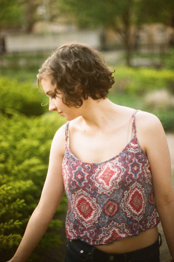 Katie looks downward, her face in profile. Her short, curly brown hair rests softly around her head, and she wears a patterned tank top that exposes her shoulders and collarbones. Behind her, low green shrubs fill the softly blurred background, with a hint of a park setting visible in the distance.