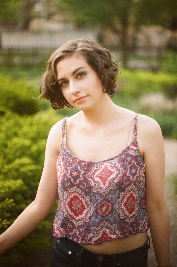 Katie turns her head slightly towards the camera while standing amidst green shrubs. Her expression is neutral but engaged, her patterned tank top detailed with geometric designs. The dreamy background features a blend of soft-focus plants and distant park elements, with warm sunlight adding a gentle glow.