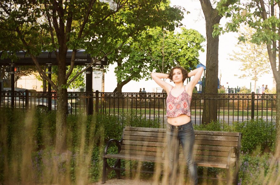 Katie is positioned mid-ground in a park-like setting. She stretches her arms behind her head while standing in front of a wooden bench, surrounded by trees and a metal fence. The foreground includes soft, out-of-focus plants, and the background displays hints of urban structures and greenery. The film captures a mix of sharp and blurred details.