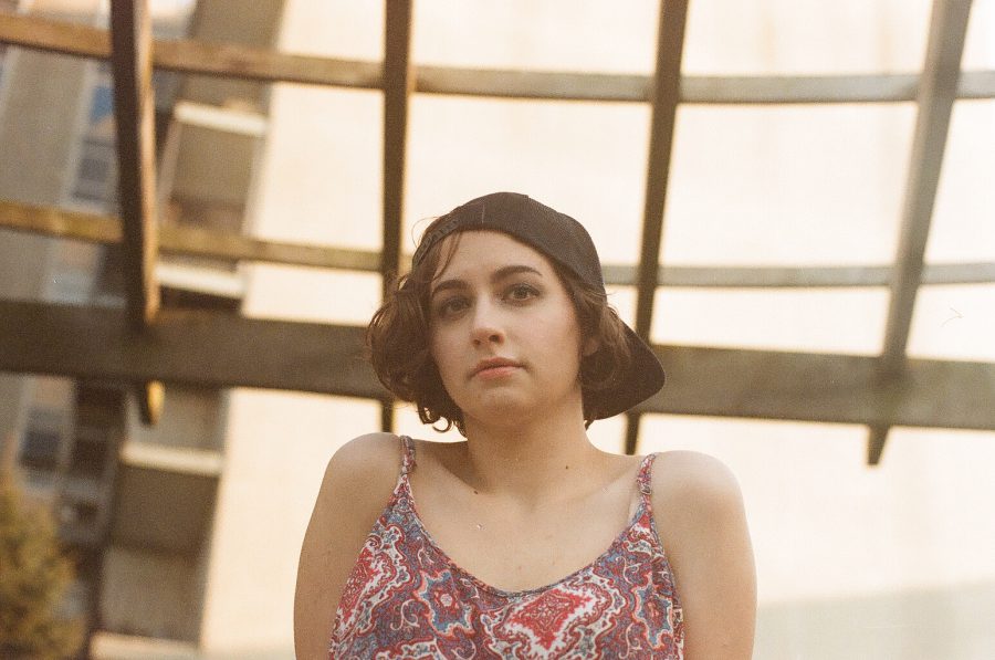A low-angle view of Katie shows her wearing a backward black cap and a geometric-patterned tank top. She looks ahead with a neutral expression while standing under a geometric wooden structure. The background features urban architecture, softened by the natural lighting and shallow depth of field.