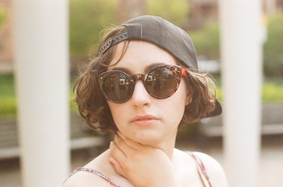Katie, wearing large sunglasses and a backward black cap, is framed close-up. Her hand rests on her neck as she gazes slightly away from the camera. Behind her, park benches and green shrubs are softly blurred, adding depth and contrast to the portrait captured on film.