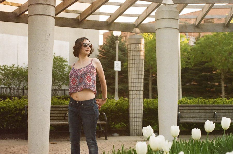 Katie stands confidently in a park surrounded by columns of a modern structure. Her patterned tank top and jeans contrast with the vibrant green bushes and a bed of white tulips in the foreground. Benches and trees populate the background, captured with a warm, natural film tone.