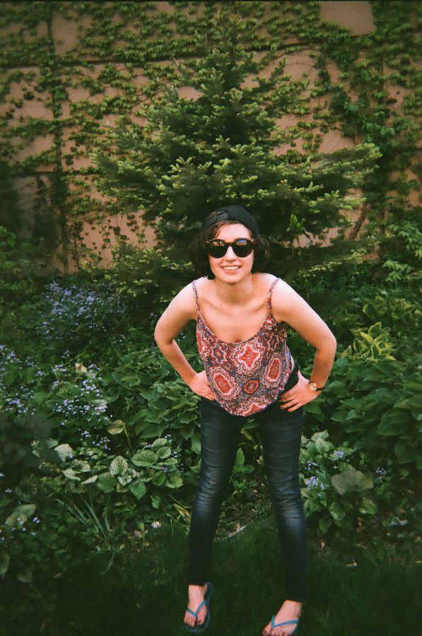 Katie leans forward playfully, her hands on her knees, while wearing sunglasses and a backward black cap. She smiles broadly in front of a background covered in dense green vines and bushes. Her patterned tank top and dark jeans are well-lit, and the soft focus of the film enhances the vibrant outdoor scene.