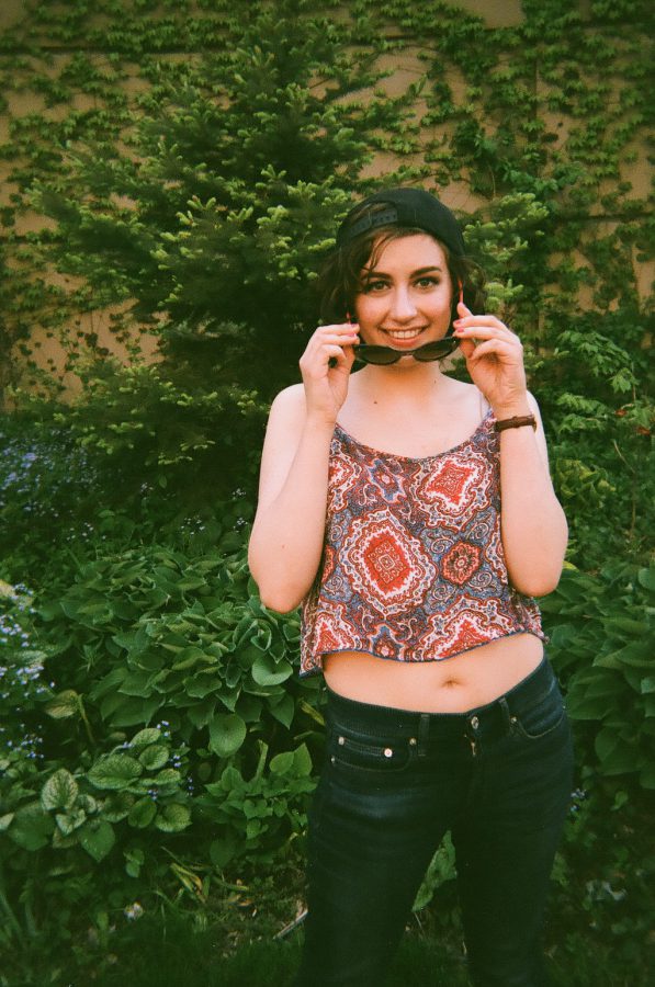 Katie stands in front of a lush background filled with green vines and bushes. She is pulling her sunglasses down to her nose with both hands while smiling at the camera. She wears a patterned tank top and a backward black cap. The film captures soft, natural light and the vibrant greenery around her.