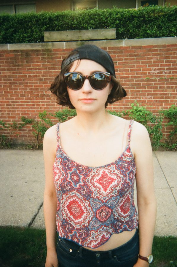 Katie stands in front of a brick wall, wearing her black cap backward and dark sunglasses. Her red and blue patterned crop top reveals a portion of her midriff, and she pairs it with dark jeans. Behind her, low bushes line the sidewalk, which leads to more greenery in the distance. The image captures her casual yet confident stance with crisp focus on her face and outfit, highlighting the textured film quality.