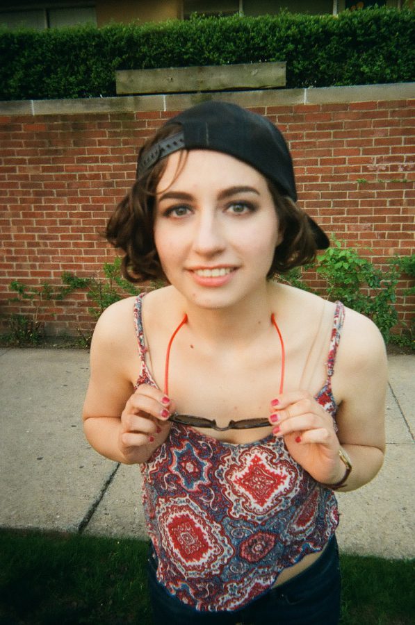Katie leans slightly forward, holding her dark sunglasses in both hands as she removes them. Her black cap remains backward on her head, and her short curly brown hair frames her smiling face. She wears the same red and blue patterned crop top and jeans, with a brick wall and greenery visible in the background. The slightly tilted angle of the film photo emphasizes her playful gesture.