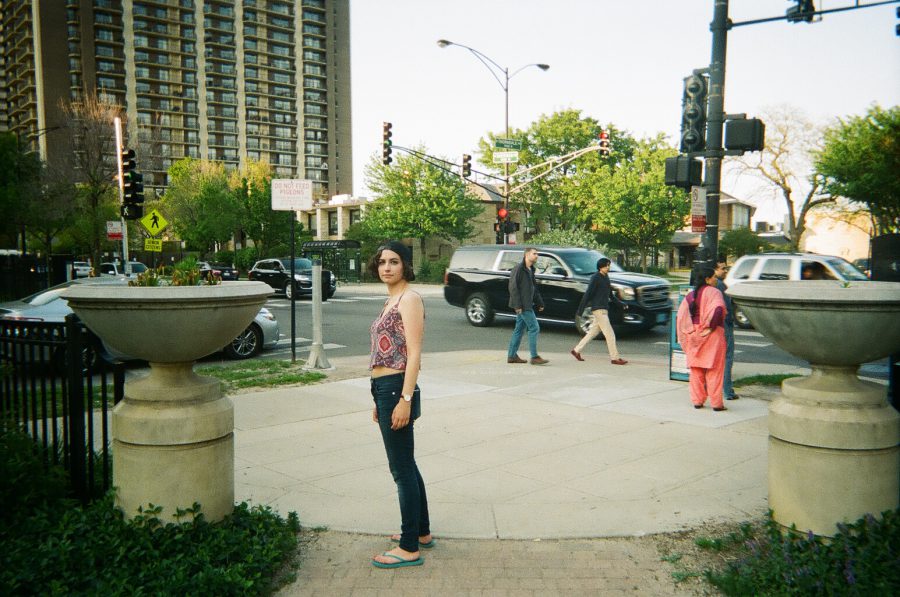 Katie poses in an open area near a busy Chicago intersection. She stands between two large cement planters, wearing her red and blue patterned crop top and dark jeans. Behind her, pedestrians cross the street, cars pass by, and tall apartment buildings line the horizon. The photograph captures the urban environment and her poised, relaxed stance.