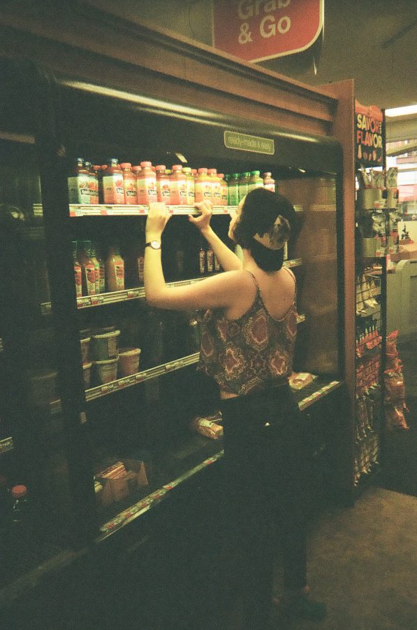 Katie is photographed indoors at a convenience store, reaching up toward the top shelf of a cooler labeled “Grab & Go.” Her black cap is backward, and she wears her red and blue patterned crop top, exposing her arms as she stretches. The dim lighting inside the store casts a warm tone over the film image, contrasting with the vibrant colors of the drink bottles on the shelves.
