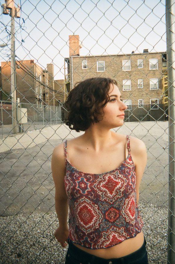 Katie stands against a chain-link fence in Chicago, with a gravel lot and older brick buildings visible in the background. She’s wearing her red and blue patterned tank top and dark jeans. Her short, curly hair frames her face as she looks off to the side, her hands relaxed behind her back.