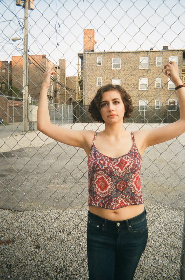 Katie faces the camera, holding onto the chain-link fence with her arms raised. Her red and blue patterned tank top and dark jeans are visible, while the gravel lot and brick buildings in the background provide a contrast to her direct, steady gaze. The photo captures her pose, emphasizing the urban surroundings.