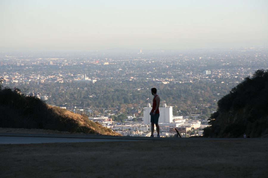 Standing on a dirt trail at Runyon Canyon, I look back over the cityscape of Los Angeles. My silhouette is framed neatly against the expansive view, with the foreground capturing the rugged trail and canyon vegetation. I wear a red sleeveless shirt and shorts. Behind me, the city stretches out endlessly, fading into a soft, atmospheric haze at the horizon, with iconic Los Angeles landmarks faintly visible in the distance.