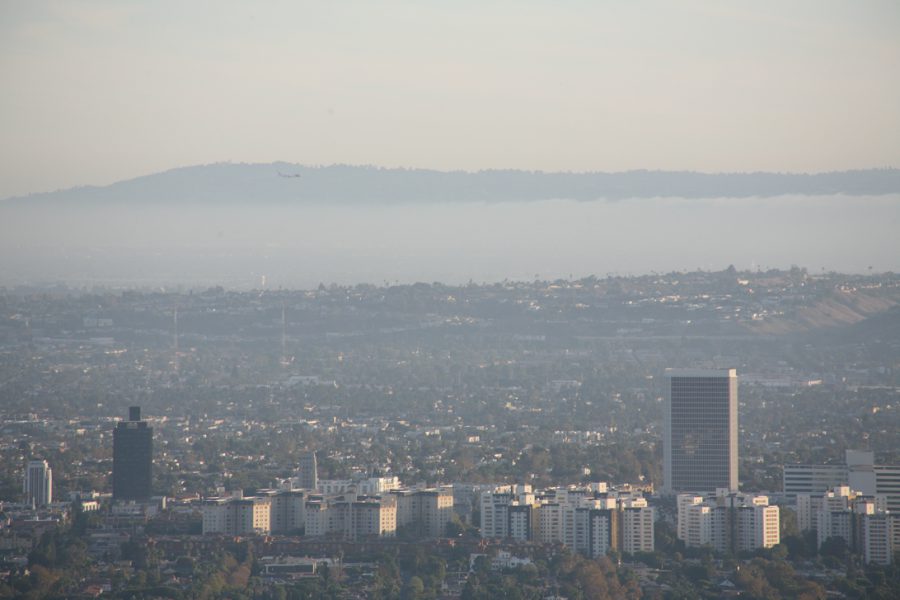 A zoomed-in view from Runyon Canyon focusing on Los Angeles, with the city skyline in the middle ground. Closer to the horizon, the distant landmass might be Catalina Island or another southern California mountain range, highlighted by a layer of atmospheric haze. The foreground is absent of significant details, allowing the focus to remain on the contrast between urban density and distant natural landscapes.