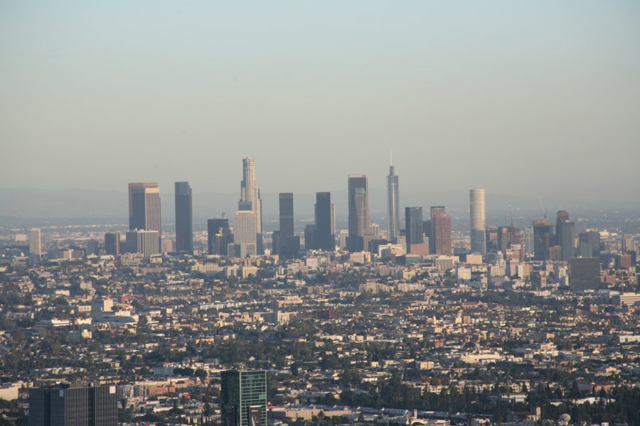 A classic downtown Los Angeles view taken from Runyon Canyon. Prominent skyscrapers, including the U.S. Bank Tower and Wilshire Grand Center, dominate the scene. The city stretches out in a dense, grid-like fashion, showcasing a mix of industrial, commercial, and residential areas. The atmosphere adds a slight haze to the horizon, softening the edges of the skyline and creating a layered effect.