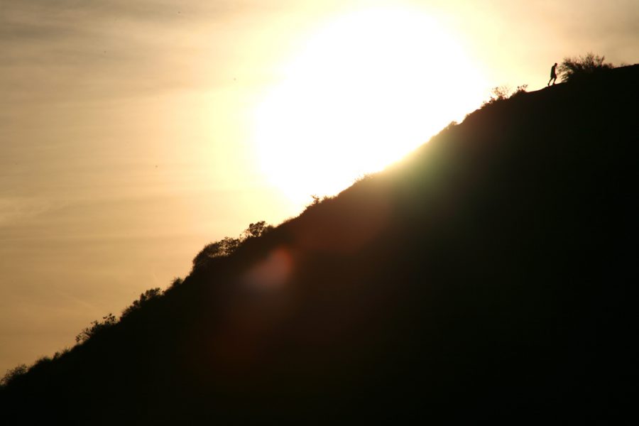 A striking sunset captured from Runyon Canyon, with a silhouetted hillside rising diagonally across the frame. A single human figure stands near the top of the hill, outlined against the radiant orange glow of the setting sun. The bushes and rugged landscape in the foreground contrast sharply with the bright, almost blinding sunlight that envelops the sky.