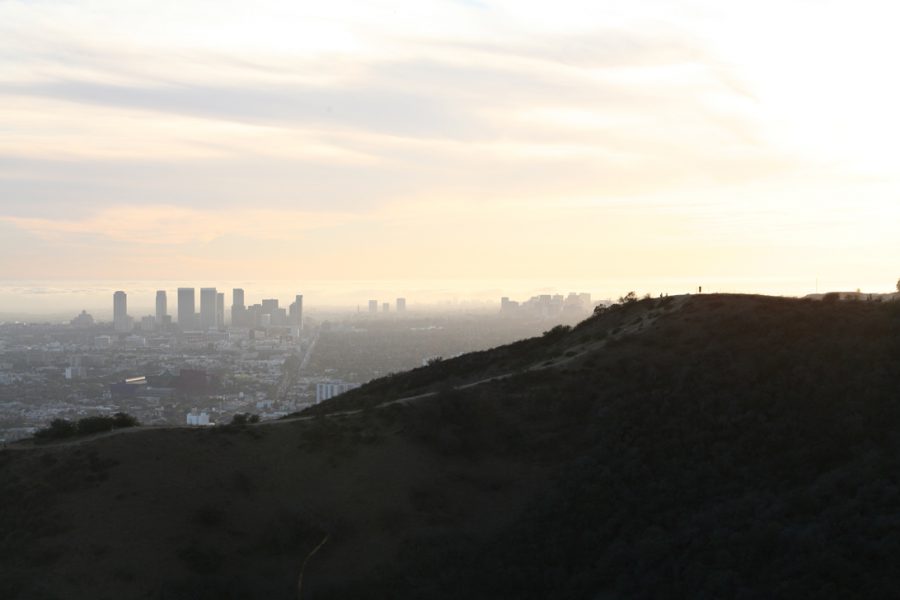 A darker, nearly silhouetted hillside view from Runyon Canyon with one or two people faintly visible at the edge of the trail. The hazy atmosphere of the Los Angeles basin fills the background, blending into the brightness of the late afternoon sky. The photo highlights the natural contours of the hill against the expansive urban sprawl, softened by the distant glow of sunlight.